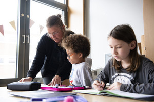 Zu sehen sind eine Lehrerin und zwei Schüler in einem Klassenraum. Die Lehrerin spricht mit dem linken Schüler.