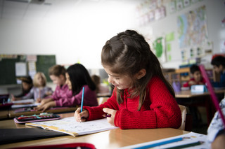 Eine Schülerin der ersten Klasse schreibt in ihr Schulheft. Im Hintergrund sind weitere Schülerinnen und Schüler zu sehen, die konzentriert arbeiten.